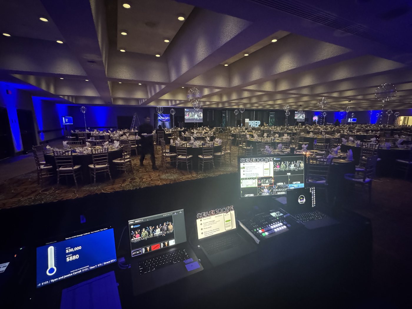 A gala ballroom seen from the production desk, live fundraising totals and event graphics running on screen