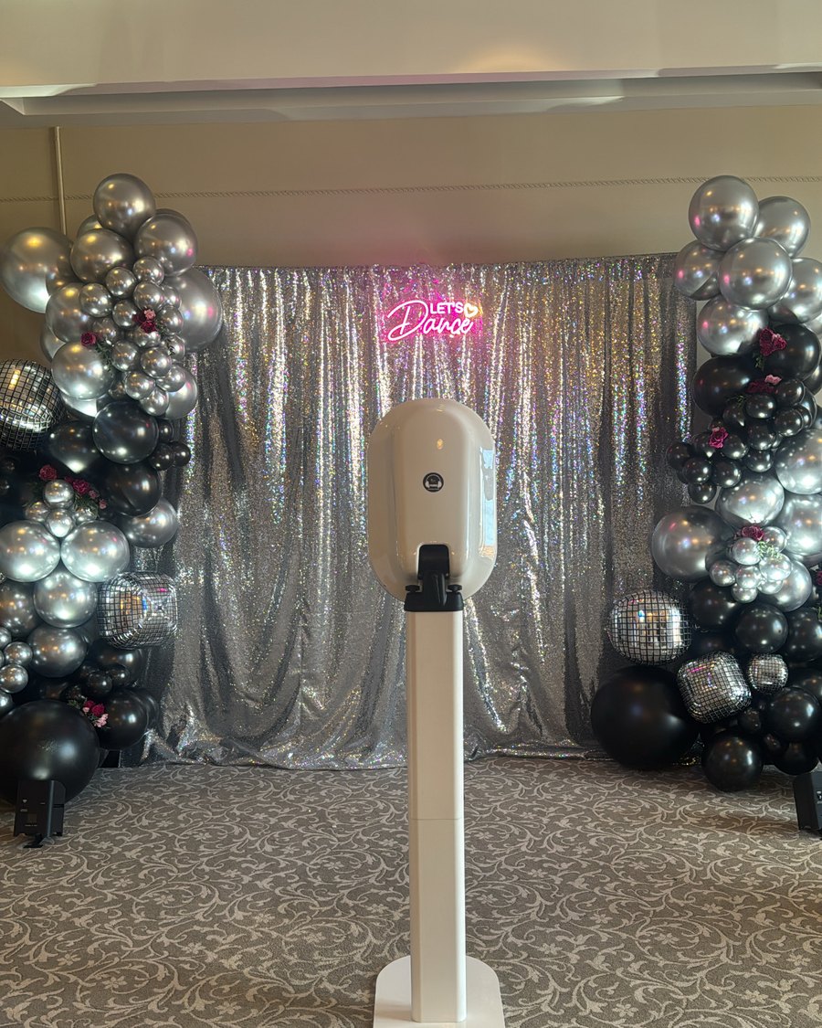A Primo photo booth in front of a silver sequin backdrop with a balloon arch and a neon Dance sign