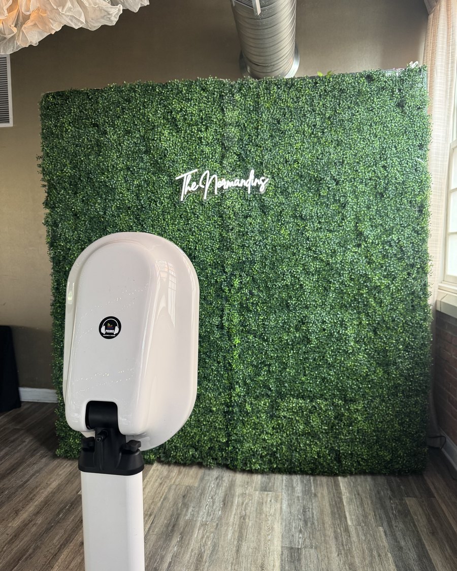 A Primo photo booth in front of a greenery wall with a custom name sign