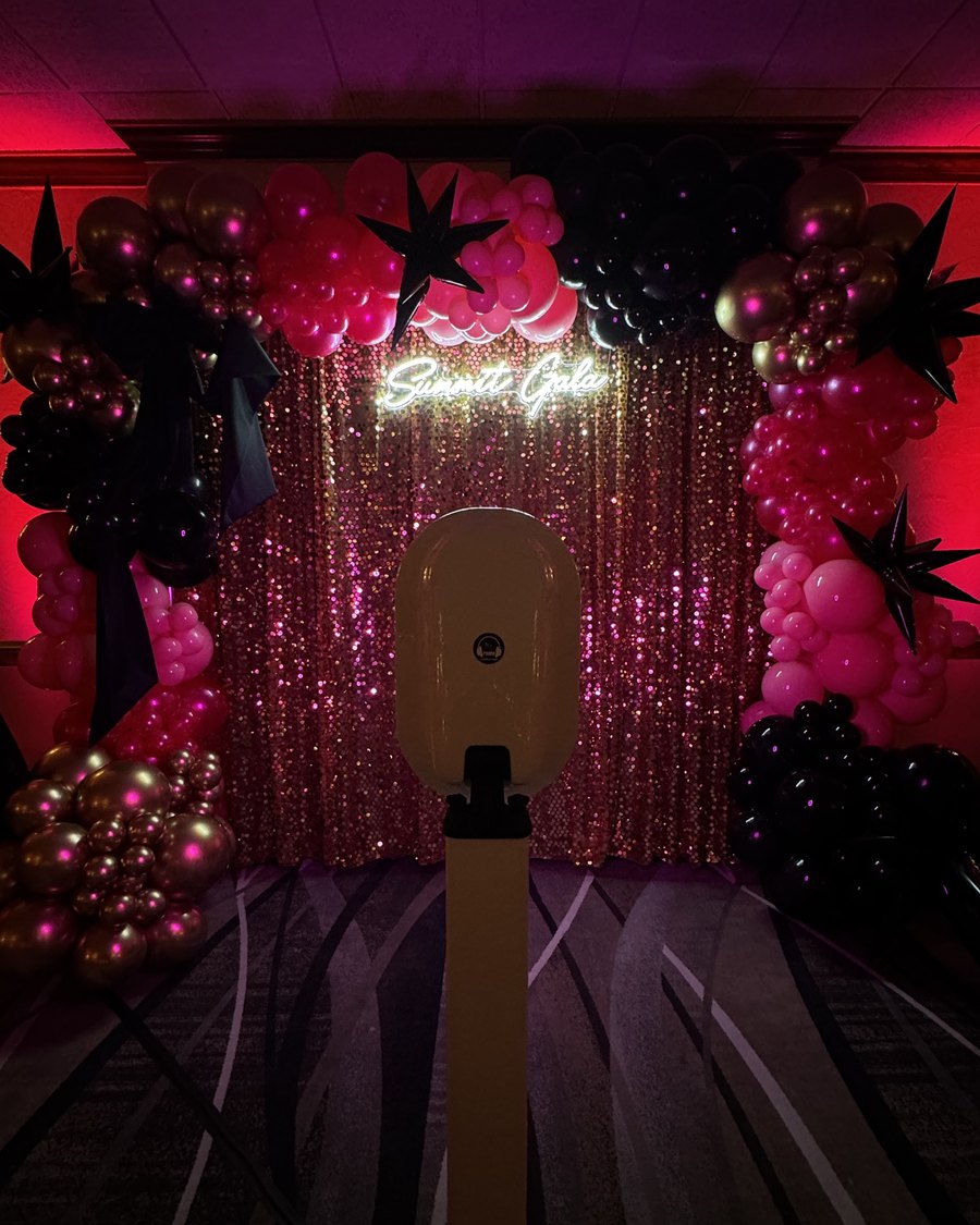 A sleek modern Primo photo booth in front of a pink sequin backdrop with a balloon arch and neon sign